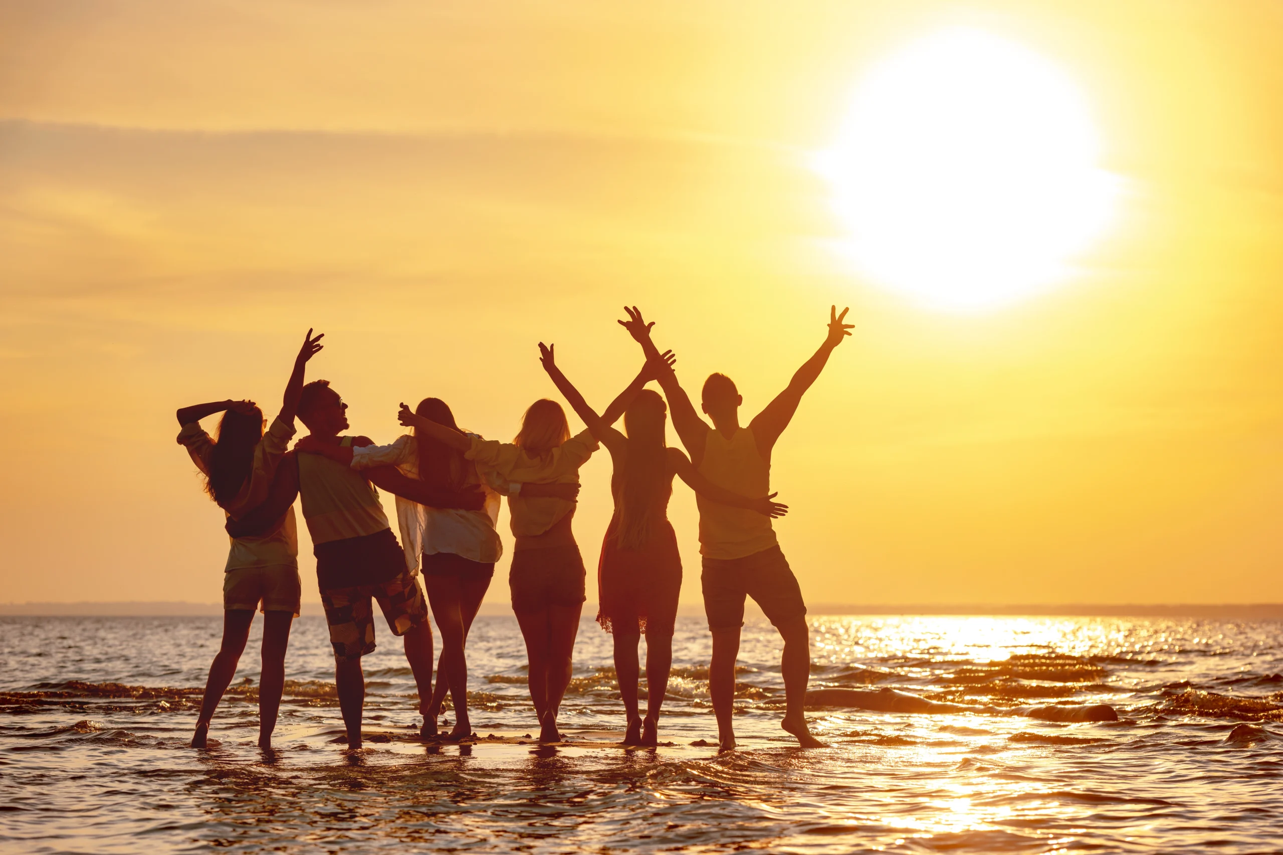 A group of friends silhouetted against a golden sunset, standing joyfully in shallow ocean water with arms raised.